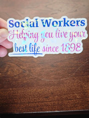Holographic sticker with text 'social workers helping you live your best life since 1898' held over a wooden surface.