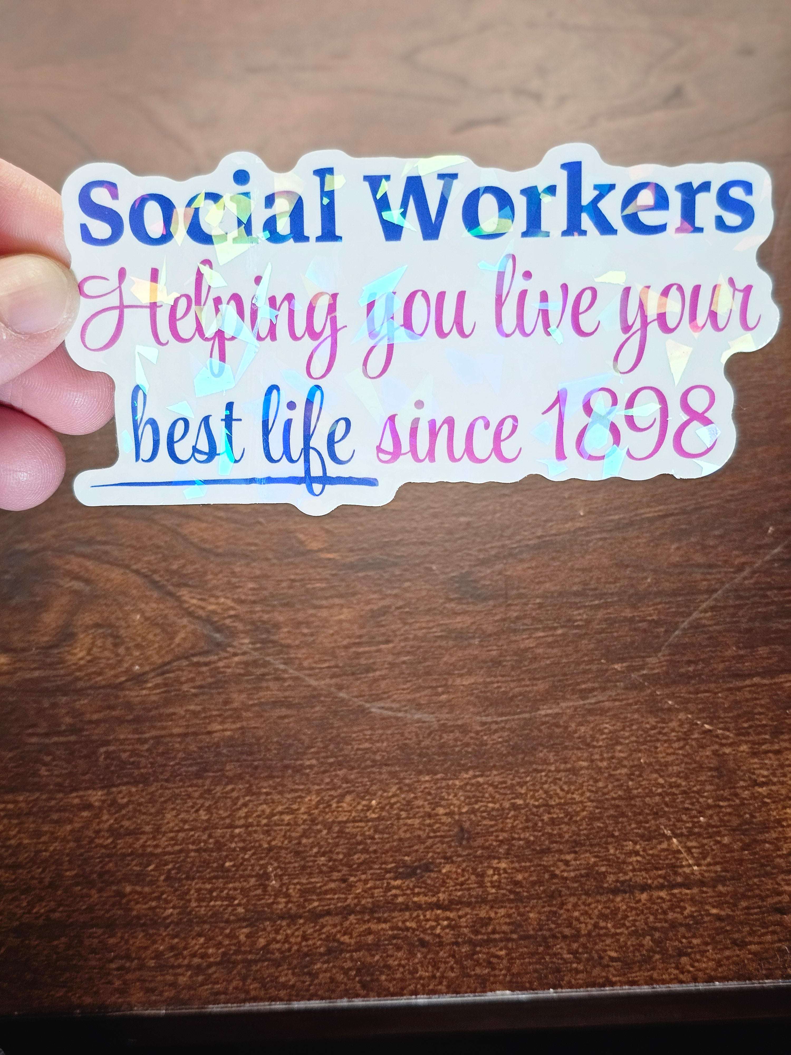 Holographic sticker with text 'social workers helping you live your best life since 1898' held over a wooden surface.