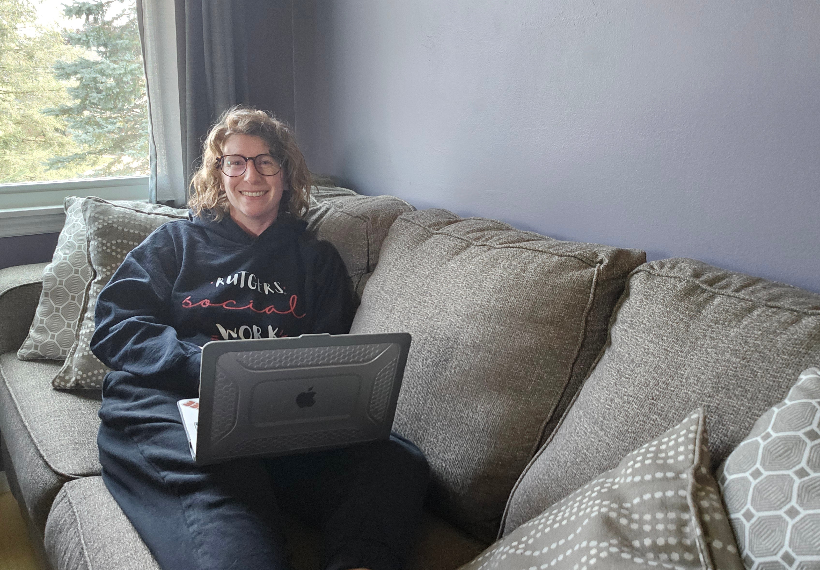 Person sitting on a couch with a laptop, wearing a dark hoodie with text, in a home setting.