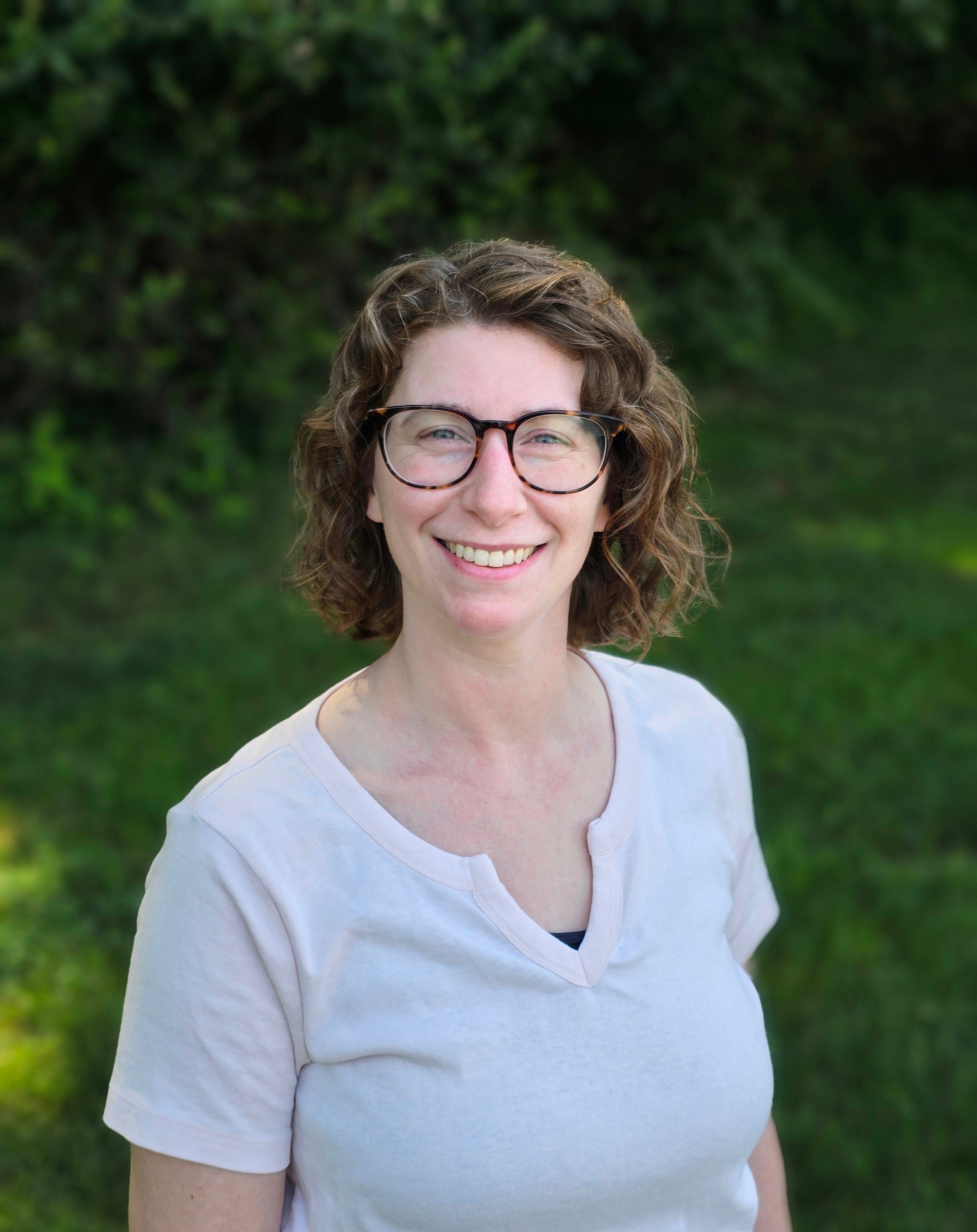 Woman wearing glasses and a white shirt standing in front of green foliage